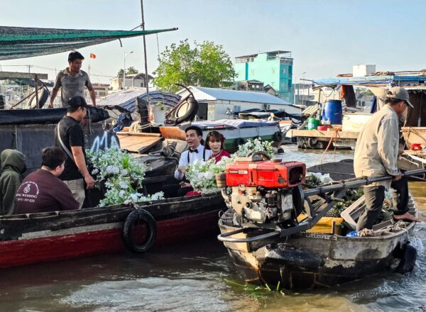 Group of people sitting and standing on boats decorated with flowers, moving through a busy floating market.