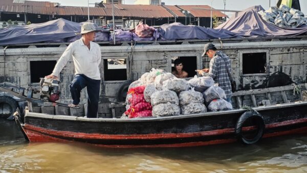 People loading and handling large bags of goods on a small boat beside a larger vessel on the river.