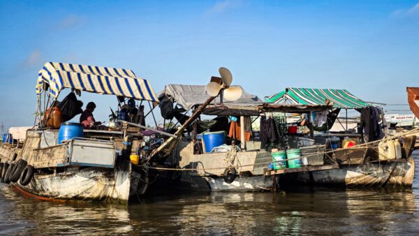 Several floating boats clustered together on a river, carrying containers, equipment, and covered living areas.