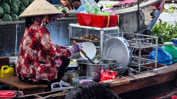 Person cooking food on a small wooden boat, surrounded by pots, dishes, baskets, and supplies on the river.