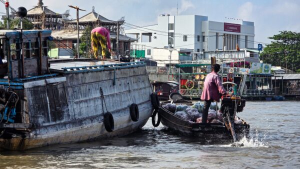 Small motorboat passing a larger weathered boat on a busy river, with people working on the boats and city buildings in the background.