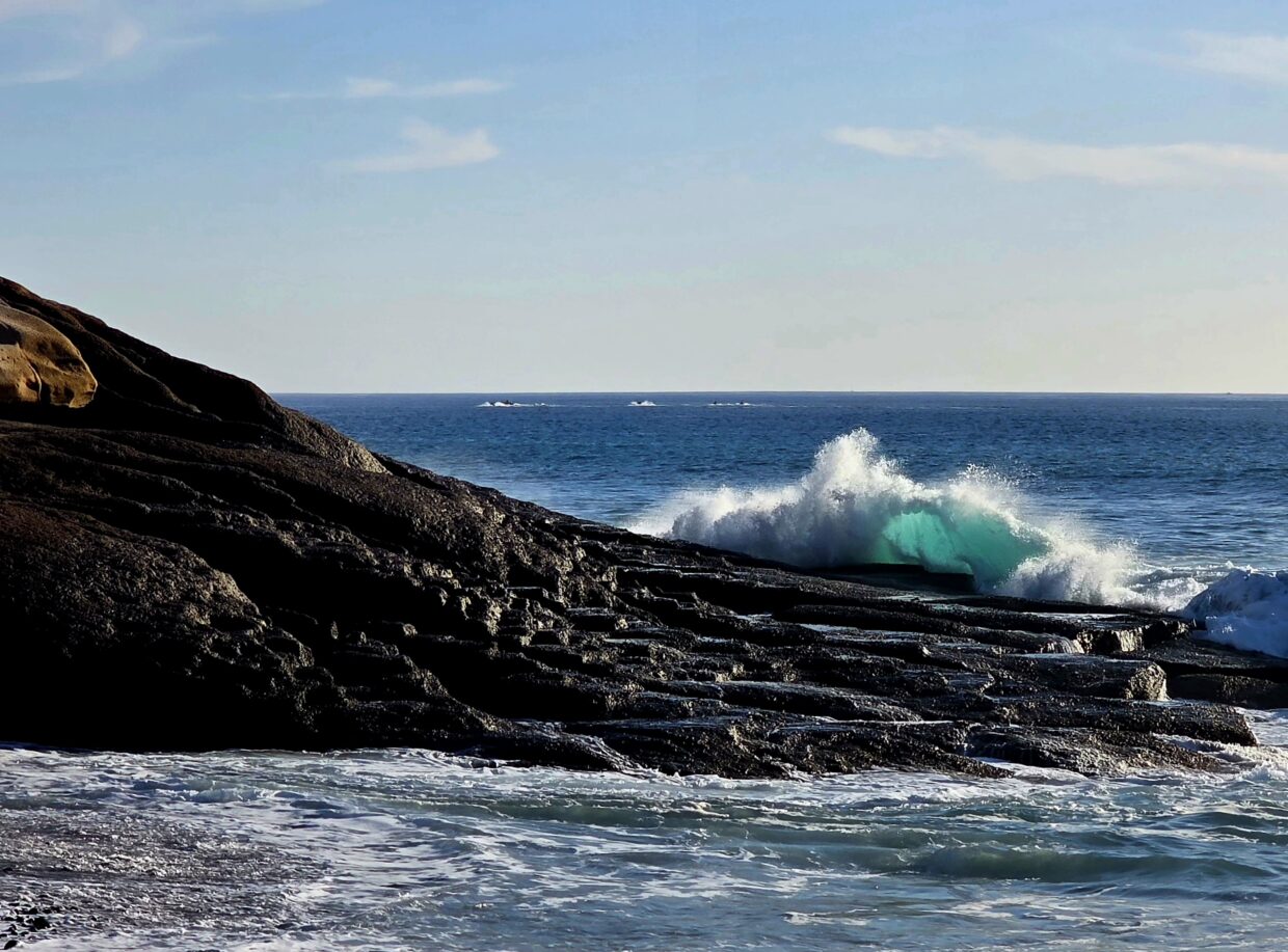 An ocean wave breaking against a dark, rocky coastline with sunlight illuminating the water in a vibrant turquoise glow.