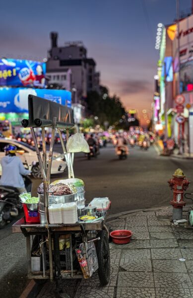 A street food cart parked on a sidewalk at a busy intersection in Saigon (Ho Chi Minh City) at dusk, with motorbikes and neon signs in the blurred background.