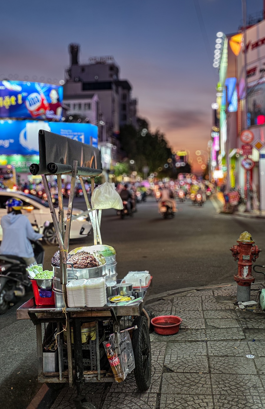 A street food cart parked on a sidewalk at a busy intersection in Saigon (Ho Chi Minh City) at dusk, with motorbikes and neon signs in the blurred background.